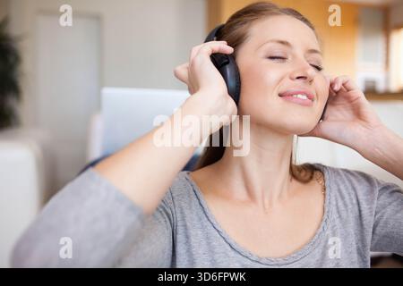 Donna seduta sul divano con cuffie sovrauricolari nere, occhi chiusi, testa inclinabile all'indietro. Donna, stile di vita, pubblicità, minimalista, interni, apertura Foto Stock