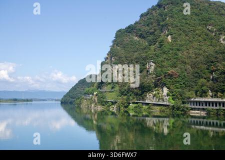 I ripidi pendii boscosi e gli affioramenti di rocce pallide si tuffano nel lago Uiam sotto Samaksan, dove una galleria protettiva si snoda lungo la riva e si riflette Foto Stock