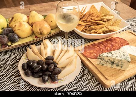 Formaggi assortiti, frutta, salumi, patatine e vino bianco disposti su un tavolo, perfetti per il cibo e lo stile di vita Foto Stock