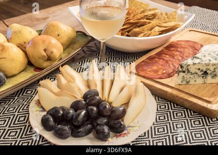 Formaggi assortiti, frutta, salumi, patatine e vino bianco disposti su un tavolo, perfetti per il cibo e lo stile di vita Foto Stock
