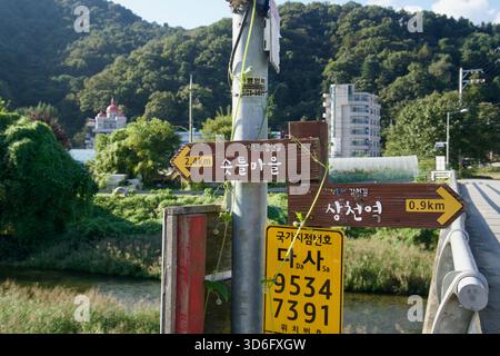 I cartelli stradali in legno indicano le indicazioni per la stazione di Sangcheon e il villaggio di Sotteul su un ponte, con un cartello giallo di localizzazione nazionale sulla posta e hi Foto Stock