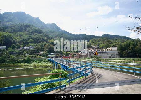 Un ponte blu porta la pista ciclabile attraverso una stretta insenatura del lago Uiam verso un piccolo villaggio, con cabine in collina e la linea della funivia visibile aga Foto Stock
