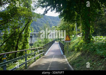 Uno stretto tratto ombreggiato della pista ciclabile di Bukhangang corre sopra il fiume con un cartello triangolare di avvertimento e vista verso la diga attraverso gli alberi. Foto Stock