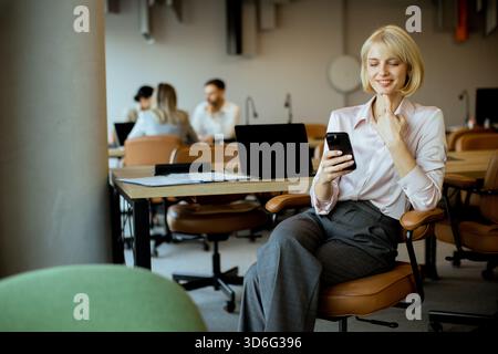 Una giovane donna dai capelli biondi siede comodamente su una sedia da ufficio, sorridendo mentre controlla il suo smartphone. Sullo sfondo, i colleghi sono impegnati Foto Stock