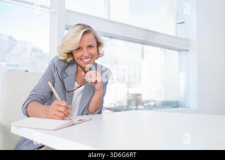Donna che indossa un blazer grigio sorridente, che tiene gli occhiali da vista e scrive su un notebook alla scrivania luminosa dell'ufficio. Professionale, executive, bionda, giacca, occhiali, Foto Stock