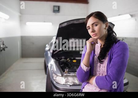 Donna a 20 anni in garage con cardigan viola, mento, cofano auto aperto, spazio per le copie. Veicolo, ispezione, contemplativo, officina, manutenzione Foto Stock