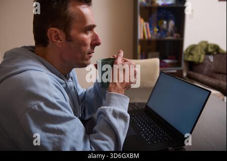Un uomo con una felpa leggera con cappuccio si siede a una scrivania, sorseggiando un caffè da una tazza mentre utilizza un computer portatile. Un ambiente tranquillo e quotidiano di lavoro remoto e vita familiare. Foto Stock