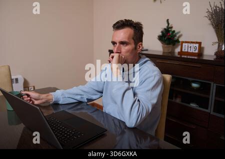 Un uomo con una felpa con cappuccio azzurra si siede a un tavolo da pranzo, concentrato e riflettente mentre lavora su un notebook a casa. Foto Stock