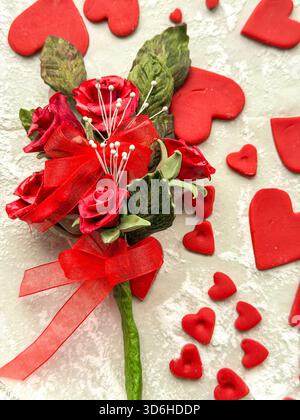 Bouquet di rose rosse artificiali con nastro circondato da forme a forma di cuore a glassa rossa sparsa su sfondo chiaro e testurizzato, artigianato da zucchero Foto Stock