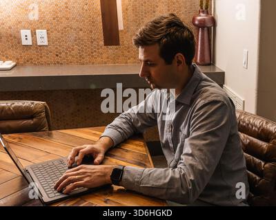 Un uomo siede su una scrivania di legno e si concentra sul proprio laptop in un ambiente accogliente. L'impostazione include un'illuminazione calda e uno sfondo a parete testurizzato, crea Foto Stock