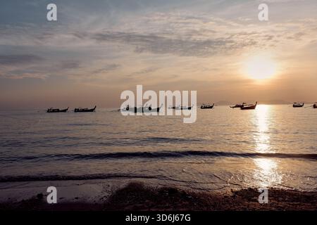 Più barche si estendono sull'acqua alla luce del mattino presto, con la luce del sole che si riflette sulle onde calme e sulle ombre tenui sulla spiaggia tranquilla. Foto Stock