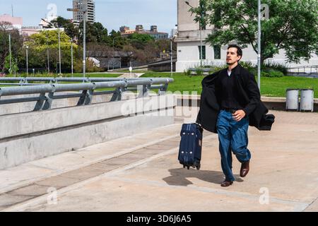 Uomo che corre e tira una valigia, si precipita a prendere un volo o un treno in un'area urbana. Stress, viaggi e concetti aziendali Foto Stock