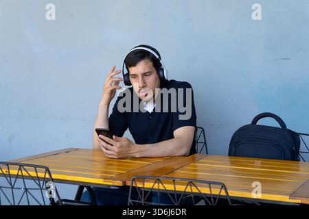 Giovane uomo che indossa le cuffie mentre usa il suo smartphone a un tavolo, sembra confuso. Lo zaino si siede sul tavolo Foto Stock