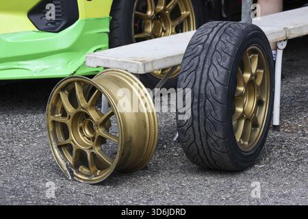 Cerchione e pneumatico in lega d'oro danneggiati sul terreno accanto alla vettura verde. parte rotta in seguito a incidente o incidente da corsa che necessita di riparazione e sostituzione del veicolo Foto Stock