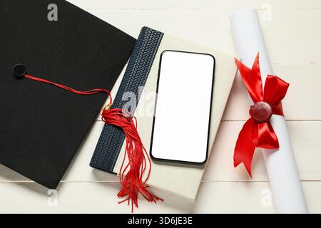 Cappello di laurea, libro, telefono cellulare e diploma su sfondo bianco in legno, vista dall'alto Foto Stock