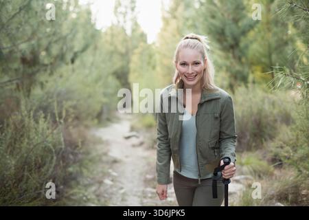 Lo stretto sentiero sterrato si snoda tra pini con un leggero filtraggio della luce del sole, palo da trekking visibile. Foresta, bosco, escursionismo, donna, all'aperto, luce solare Foto Stock