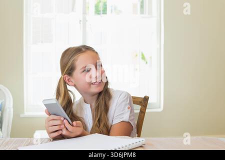 Ragazza preadolescente in camicetta bianca seduta a un tavolo di legno vicino alla finestra che sorride il telefono e il notebook. Bambino, luce solare, apprendimento, diario, casa, luce del giorno, c Foto Stock