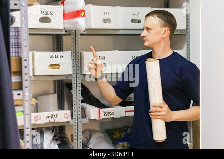 Profilo laterale MAN in alto blu che punta il dito sulle scatole bianche sul ripiano, tenendo il rotolo sotto il braccio. Foto Stock