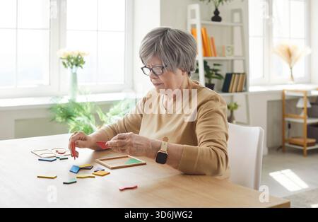 Anziana donna dai capelli grigi che raccoglie figure geometriche in legno a casa seduta al tavolo Foto Stock