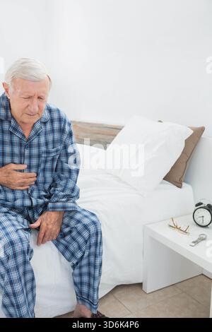 Un anziano che stringe il petto seduto sul letto indossando pigiami a quadri blu accanto alla sveglia, spazio fotocopie. Anziani, uomini, camera da letto, orologio, occhiali, di Foto Stock