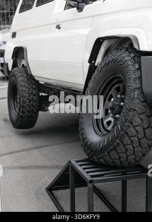 Vista ad angolo basso di un SUV fuoristrada bianco sollevato su una rampa di esposizione, evidenziandone l'articolazione. Lato di un 4x4 personalizzato con pneumatici fuoristrada aggressivi Foto Stock