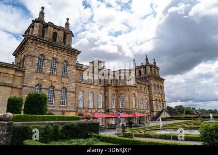 Woodstock, Regno Unito, 12 luglio 2023, Golden Stone Building con Twin Towers e finestre ad arco. Ombrelloni rossi e fontane del giardino sotto una nuvolosa Foto Stock