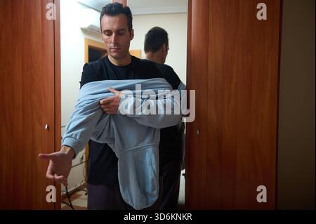 Un uomo si trova su una porta di legno, sollevando una felpa con cappuccio blu mentre indossa una camicia scura, con un aspetto premuroso. Ideale per la vita a casa, la famiglia, il lavoro e tutto il resto Foto Stock