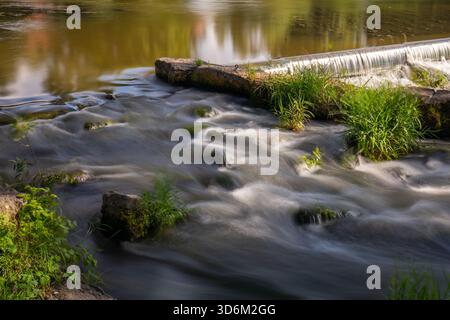 Una fotografia tranquilla e a lunga esposizione cattura il morbido e setoso flusso d'acqua che si getta su una piccola diga di pietra artificiale o una diga su un fiume. La mozione Foto Stock