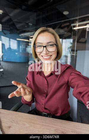 Donna d'affari felice che indossa occhiali da vista e una camicia marrone, sorridente e gestuale mentre partecipa a una riunione virtuale o a una conferenza online dal suo spazio di lavoro aziendale Foto Stock
