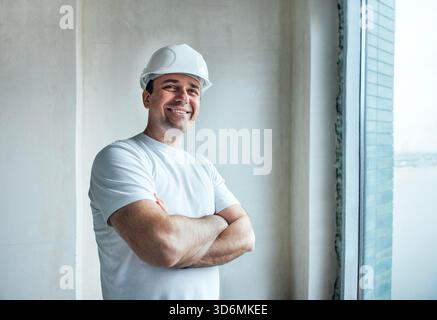 Il lavoratore edile con elmetto si erge con sicurezza con le braccia incrociate, evidenziando uno spazio interno luminoso in fase di costruzione con grandi finestre e pareti non finite || modello rilasciato Foto Stock