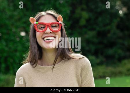 Donna sorridente mentre è in piedi all'aperto in giardino con un maglione in maglia beige e occhiali rossi Foto Stock