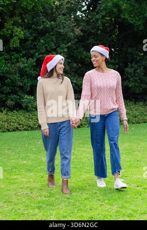 Diverse amiche che camminano mano nella mano attraverso il prato in giardino con i cappelli di Babbo Natale Foto Stock
