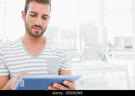 Uomo con barba che indossa una T-shirt a righe utilizzando un tablet azzurro su scrivanie bianche e monitor in ufficio. Spazio di lavoro, dispositivo, schermo, moderno, luminoso, urbano, skyli Foto Stock