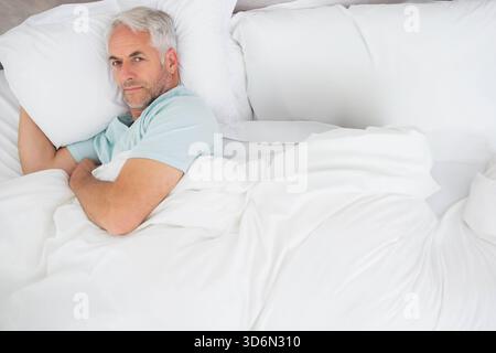 Uomo anziano sdraiato sul letto con maglietta azzurra in mezzo a piumone e cuscini bianchi, spazio fotocopie. Anziani, riposo, calma, serena, minimalista, neutrale, a letto Foto Stock
