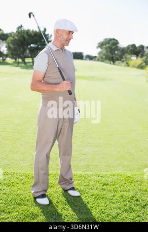 Uomo maturo in piedi sul fairway del golf che tiene il ferro da golf sulla spalla indossando un cappello piatto e un guanto. Golfista, all'aperto, illuminato dal sole, club, focus, serenità, natura Foto Stock