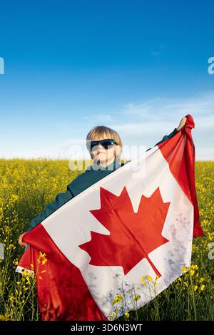 Ragazzo con una grande bandiera canadese contro un campo di canola giallo in una giornata di sole. Buon Canada Day. Orgoglio per il paese, la libertà, il patriottismo. Diaspora, immigr Foto Stock