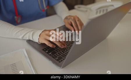 Infermiera donna con stetoscopio che scrive su un computer portatile con mani tatuate in un moderno edificio clinico; concentratevi sulla dedizione. Foto Stock
