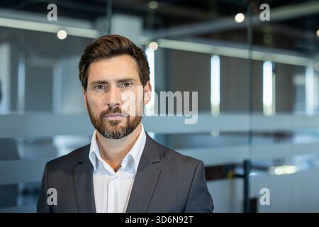 Uomo d'affari sicuro in un moderno ufficio in vetro, indossa un abito e guarda direttamente la fotocamera con un'espressione seria e determinata che trasmette leadership e successo aziendale Foto Stock