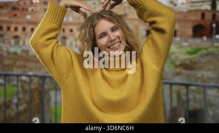 Una donna solleva le braccia a forma di cuore vicino al panorama di roma mentre un giovane turista biondo emette un sorriso caldo indossando un comodo maglione giallo mentre ammira il paesaggio Foto Stock