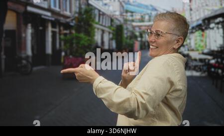 La donna punta il dito in avanti sulla strada pedonale mentre sorride e tocca il petto con l'altra mano, i capelli corti e gli occhiali sono visibili; gioia giocosa. Foto Stock
