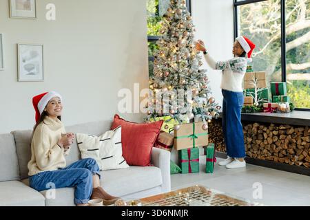 Diverse amiche che indossano cappelli di Babbo Natale che decorano l'albero di Natale a casa tenendo in mano la tazza. Festività, festività, celebrazione, calore, compagnia, h Foto Stock