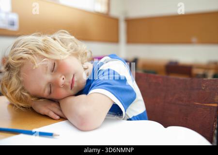 Bambino che dorme sulle braccia piegate su una scrivania in legno vicino al notebook aperto, matita blu in classe. Pisolino, allievo, tavolo, luce del giorno, calore, bokeh, calma Foto Stock