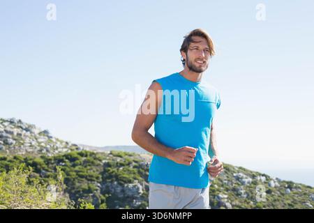 Uomo a metà degli anni Venti che corre lungo un sentiero costiero roccioso in collina con una cima senza maniche blu brillante. Atleta, jogger, all'aperto, fitness, vista mare, costa Foto Stock