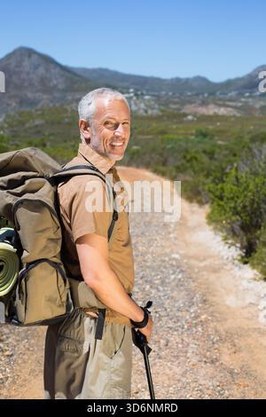 Uomo anziano in piedi sul sentiero di montagna guardando indietro, zaino di olive, pattino arrotolato legato, palo di tenuta. Escursionista, zaino, robusto, arido, paesaggio, viaggiare, o Foto Stock