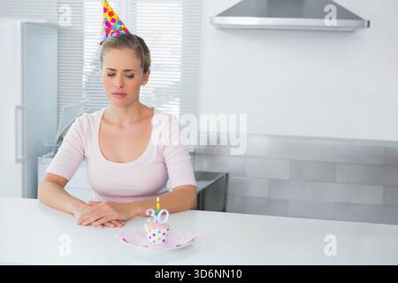 Donna di mezza età che indossa un cappello da festa, seduta al bancone della cucina, che guarda il cupcake con la luce 30. Minimalista, luminoso, frigorifero, inossidabile, luce, contemp Foto Stock