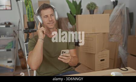 L'uomo che indossa le cuffie tiene lo smartphone e tocca le cuffie in casa tra le scatole in movimento; relax durante la giornata in movimento. Foto Stock
