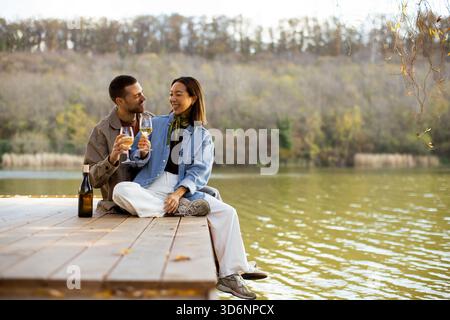 Una coppia gioiosa si siede vicino al tranquillo lago, ridendo e brindando con i loro bicchieri di vino, immergendosi in una bella giornata insieme. Foto Stock