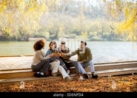 Quattro amici si riuniscono su un ponte di legno vicino al lago, allietando con le bevande mentre le foglie colorate cadono intorno a loro. Foto Stock