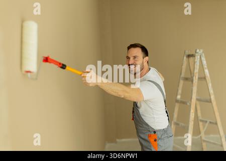 Un uomo sta dipingendo un muro con un rullo, sorridendo e godendo il processo. Indossa un vestito grigio e si trova su una superficie piana in una stanza ben illuminata Foto Stock