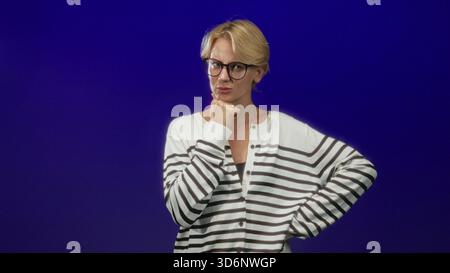 Donna con mano sul mento e palmo sul petto che indossa cardigan a righe in studio blu; contemplazione dubbio. Foto Stock